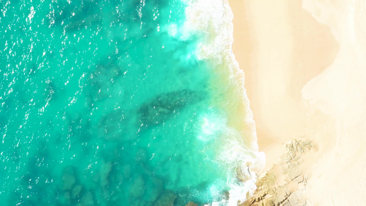 Overhead view of a turquoise wave breaking onto the sandy Baja California Sur shore, with foam curling along the beach and no visible people.