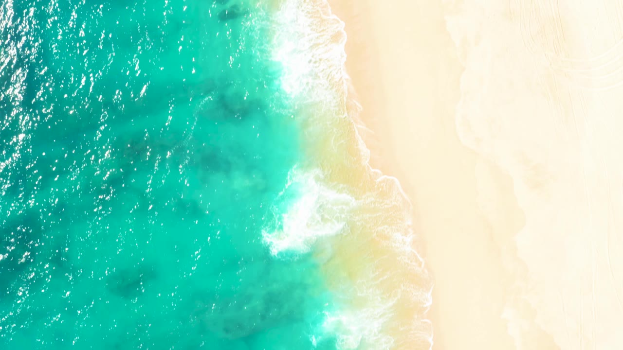 Aerial overhead frame of East Cape turquoise ocean on the left and pale beach on the right, with white foam curling at the waterline and no visible people.