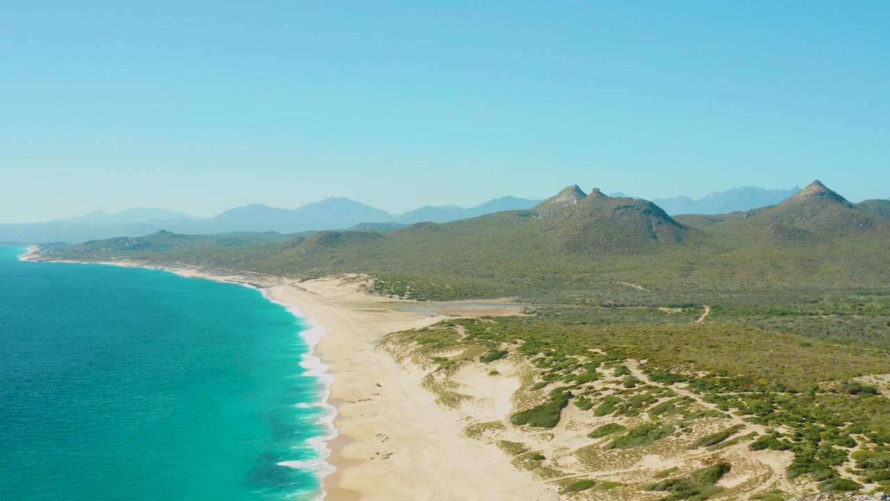 Opening frame over East Cape, Baja California Sur, with turquoise water on the left, a pale beach curving inland, dune scrub on the right, distant green mountains, clear sky, and no visible people.