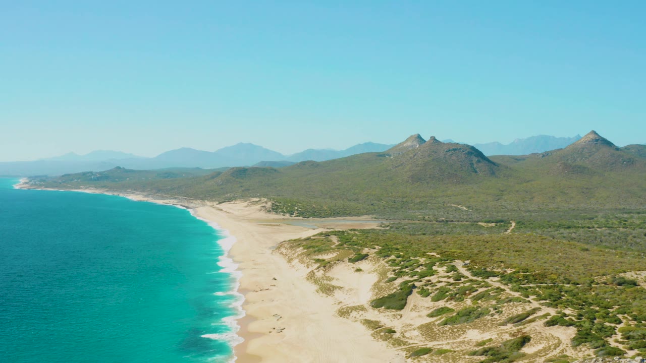 Aerial frame from East Cape, Baja California Sur, showing the shoreline slightly farther along the beach, with surf bands on the sand, scrub-covered dunes, inland hills, mountain peaks, clear sky, and no visible people.
