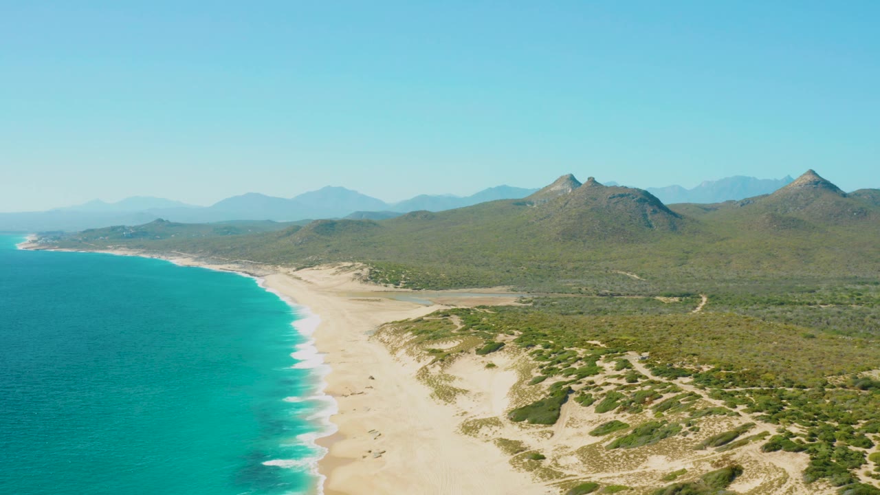 Mid-clip aerial over East Cape, Baja California Sur, with turquoise water and foamy wave edges at left, broad sand through the center, low dune plants at right, green hills beyond, clear sky, and no visible people.