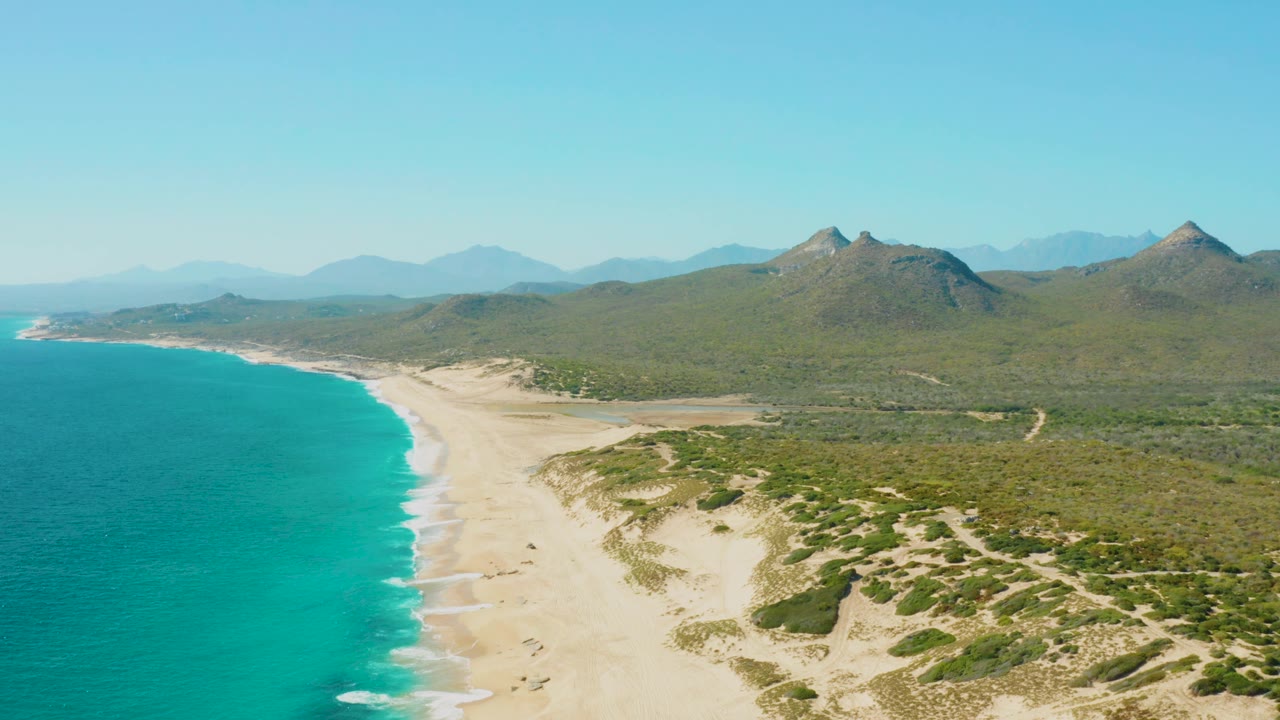 Second aerial frame of the East Cape beach in Baja California Sur, Mexico, showing the shoreline curving away, small surf bands along the sand, green inland terrain, distant mountains, and no visible people.