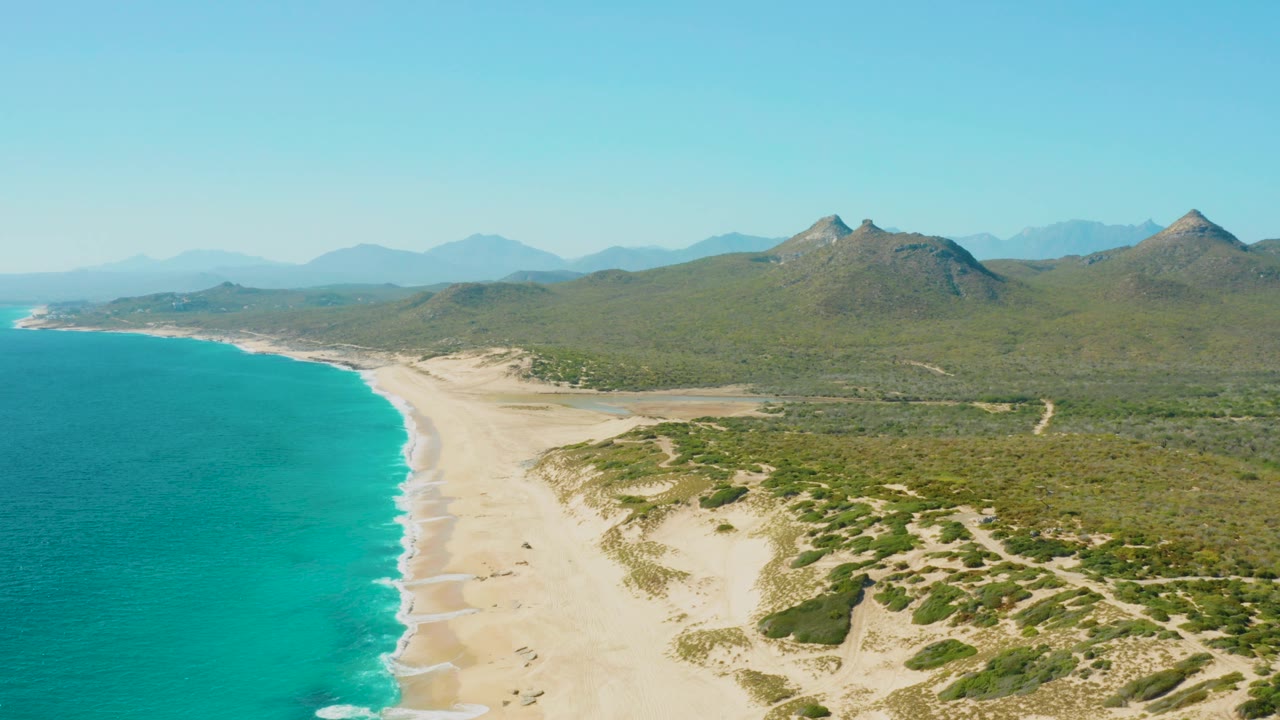 Mid-clip aerial frame over the East Cape coast in Baja California Sur, Mexico, where the water sits left, the beach runs diagonally through the frame, dune tracks and scrub are visible inland, and no visible people appear.