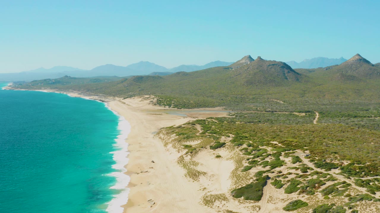 Opening aerial frame over East Cape, Baja California Sur, with turquoise water at left, a pale sandy beach, green dunes, distant mountains, clear sky, and no visible people.