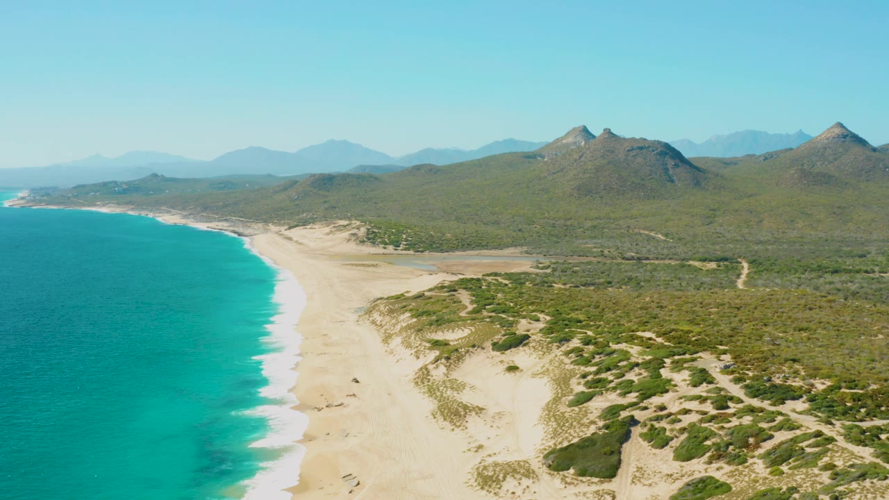East Cape aerial frame with turquoise sea filling the left side, a long pale beach through the center, scattered dune shrubs to the right, mountains on the horizon, and no visible people.