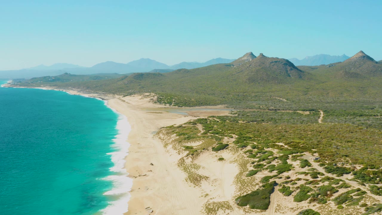 High coastal view with ocean, empty sand, dunes, and mountain peaks.
