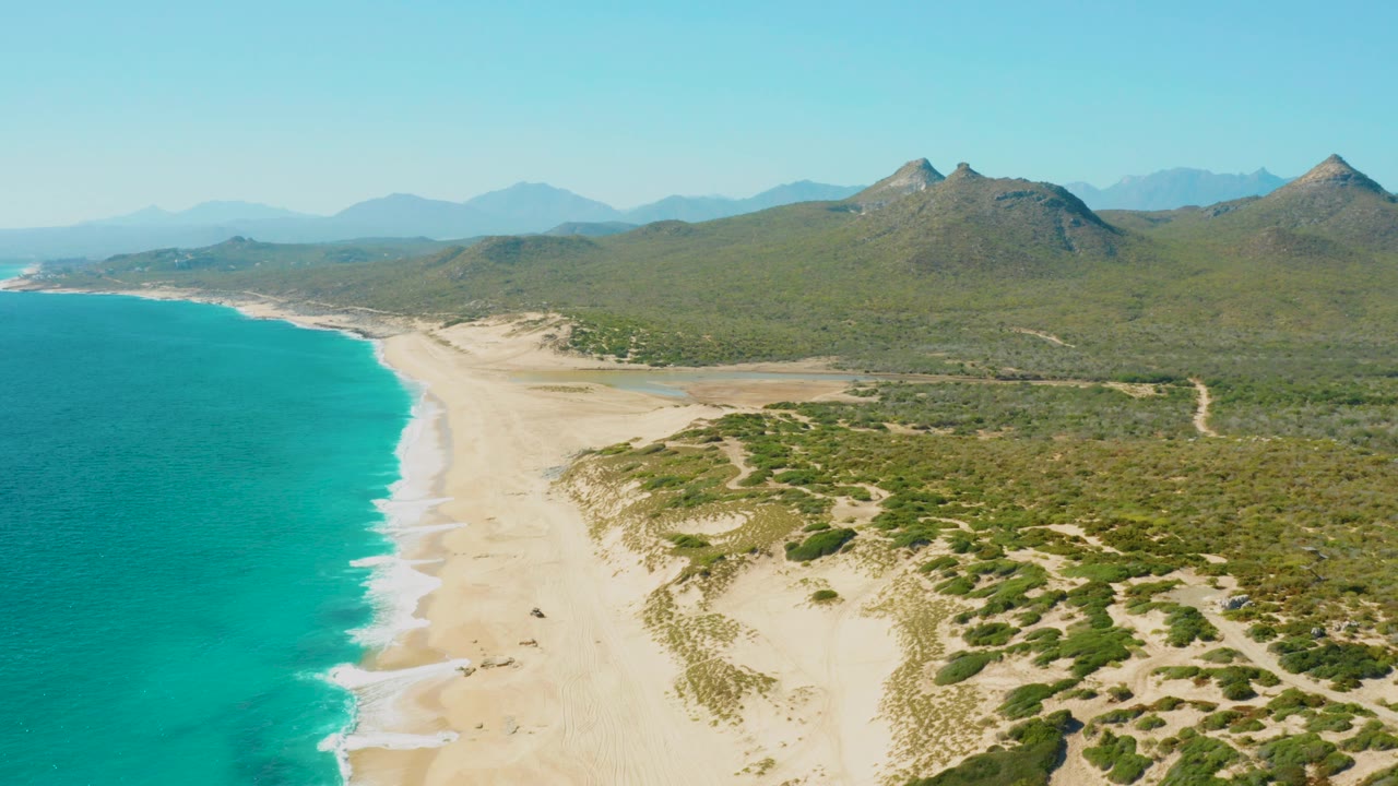 Aerial view over East Cape Baja California Sur with turquoise ocean along the left edge, pale sandy beach, green dune scrub, distant mountains, clear sky, and no visible people.