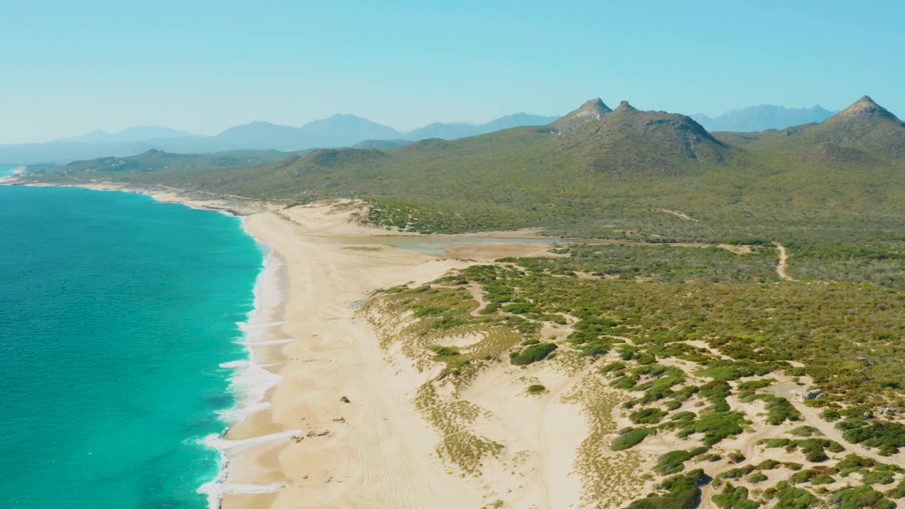 East Cape Baja California Sur coastline with turquoise ocean filling the left side, a long pale beach, low green dunes, faint inland tracks, distant mountains, and no visible people.