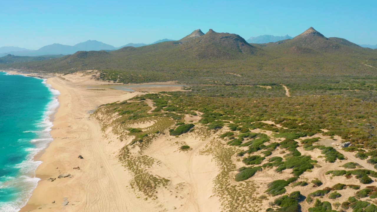 Drone view over East Cape shoreline with the surf line running up the left side, dune vegetation and sandy tracks below, a broad green plain inland, mountains on the horizon, and no visible people.