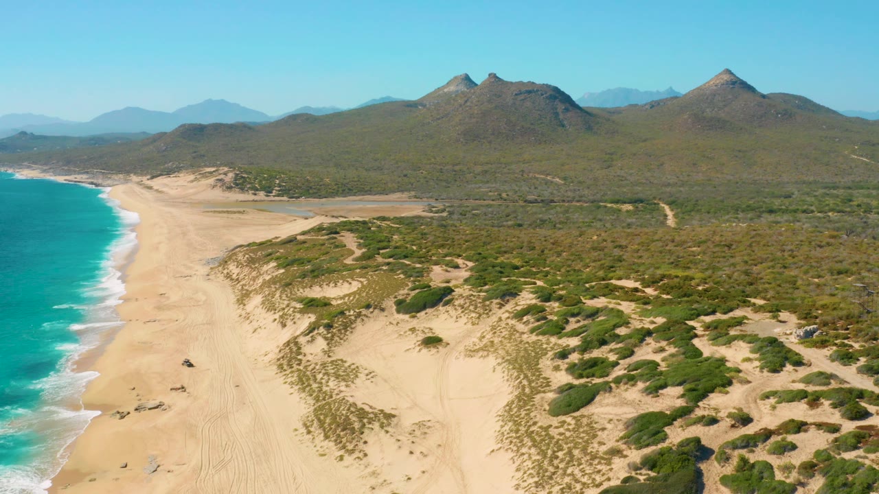 Wide East Cape aerial frame with a curving strip of turquoise ocean, light sand beach, low dunes with green shrubs, inland scrubland, distant peaks, and no visible people.