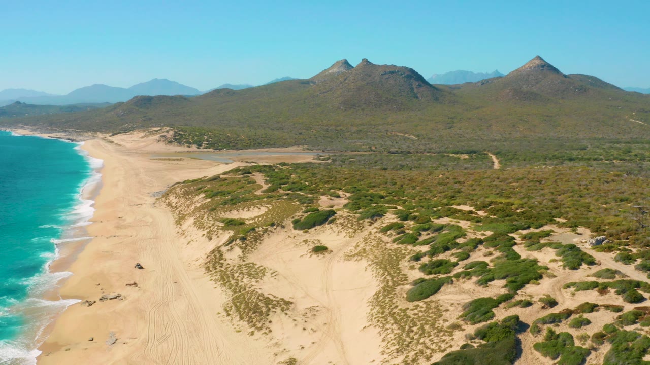 Aerial view along the Baja California Sur beach where white waves meet the sand at left, clustered dune shrubs fill the lower right, inland vegetation stretches to mountains, and no visible people appear.
