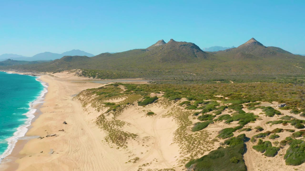 East Cape, Baja California Sur aerial view with the shoreline running diagonally, pale sand dunes in the foreground, turquoise water at left, mountain ridges beyond, and no visible people.