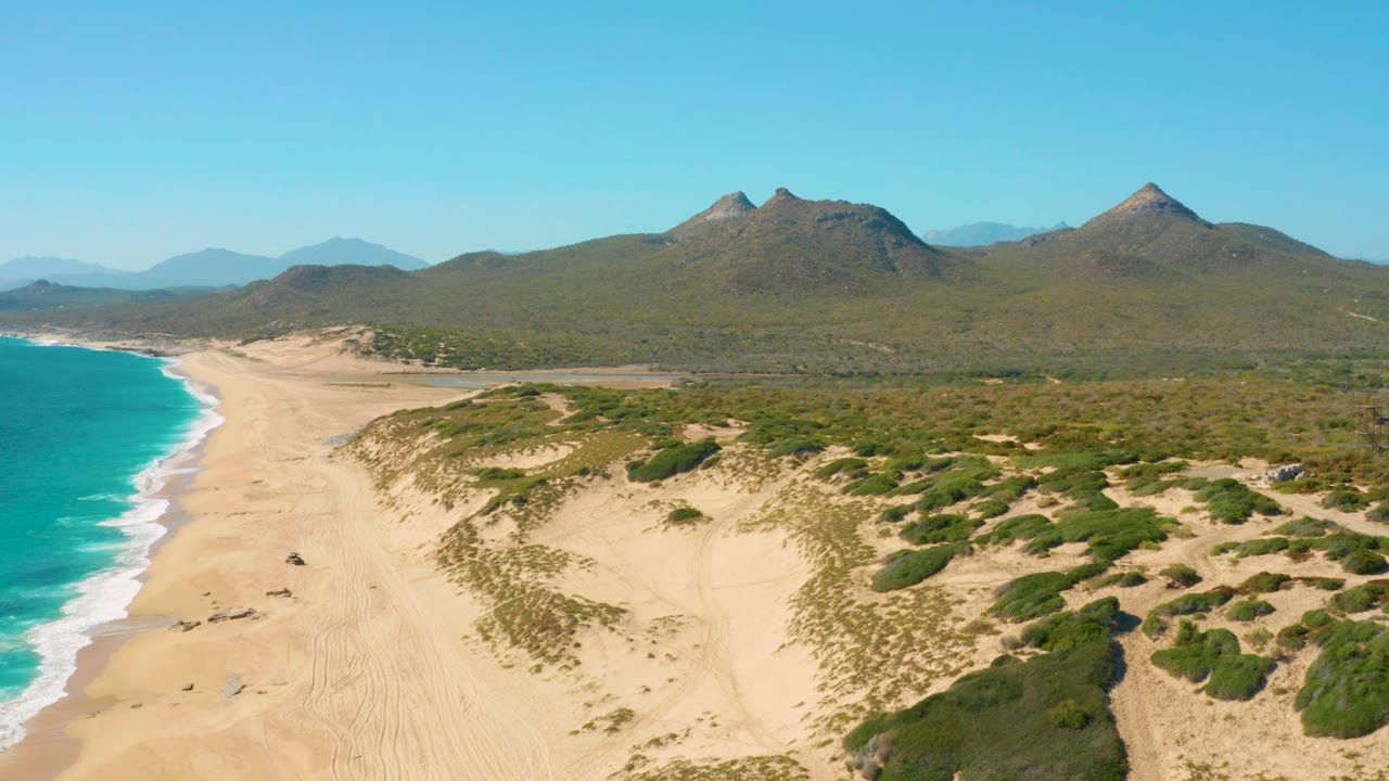 Aerial view over East Cape dunes with turquoise surf along the left edge, open sandy beach through the center, green scrub patches on the dunes, distant mountains, and no visible people.