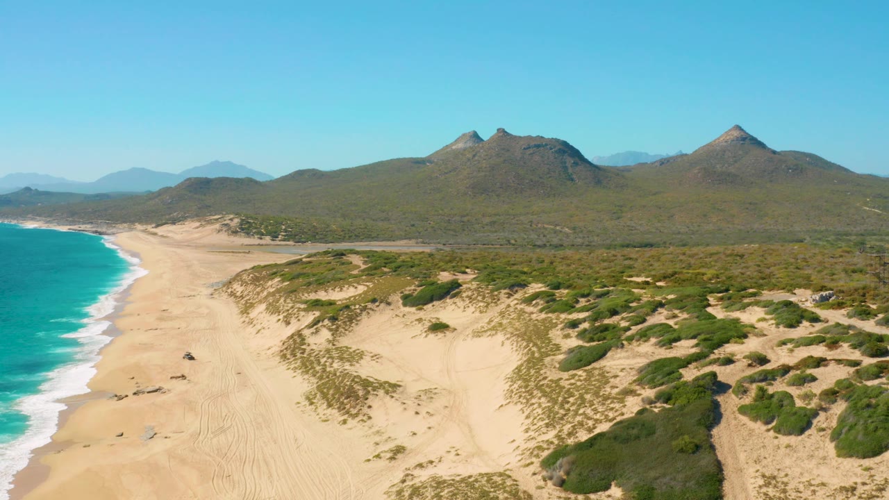 East Cape, Baja California Sur coastline from a high drone angle, with blue-green ocean and white foam on the left, wind-shaped sand and dune vegetation on the right, mountains behind, and no visible people.