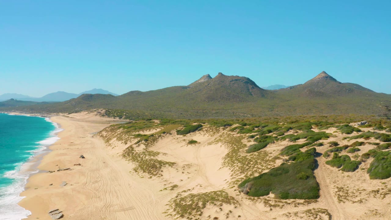 Aerial frame over East Cape dunes with the shoreline curving along the left edge, low wave foam on the sand, mountain ridges across the background, and no visible people.
