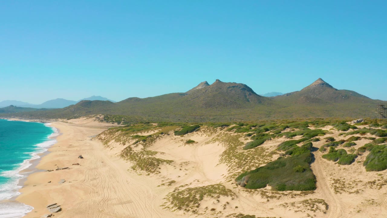 Mid-clip aerial view in Baja California Sur, with turquoise water pressing into the left shoreline, rippled sand and shrub-covered dunes ahead, mountains behind, and no visible people.