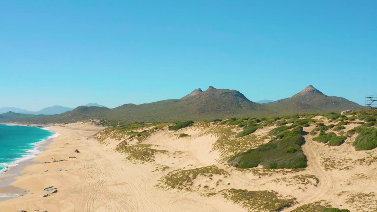 East Cape, Baja California Sur aerial view with the car farther along the open sand, surf curling at the left edge, dune vegetation across the middle ground, mountains behind, and no visible people.