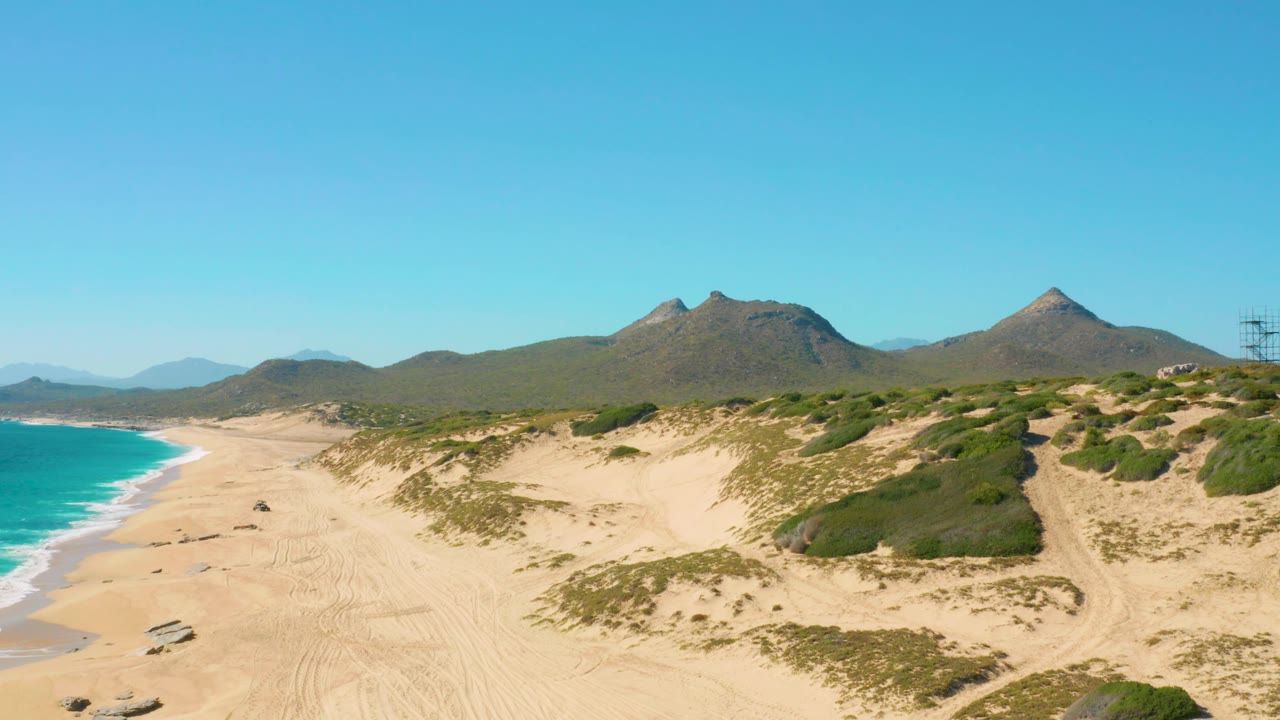 Aerial frame from East Cape, Baja California Sur, with the vehicle continuing across pale beach sand, ocean waves to the left, green dune patches to the right, distant mountains, and no visible people.