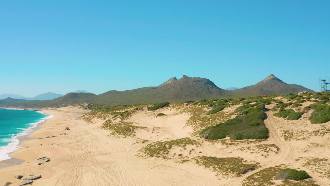 Mid-clip aerial view over East Cape, Baja California Sur, showing sand tracks leading across the beach, the small car near the shoreline side, low green dunes, dark mountains, and no visible people.