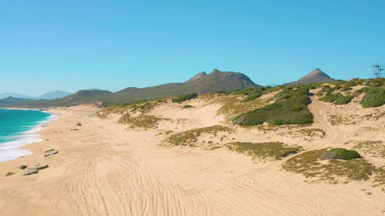 Aerial beach frame in East Cape, Baja California Sur with the vehicle slightly along the sand, ocean surf on the left edge, dune scrub to the right, and no visible people.
