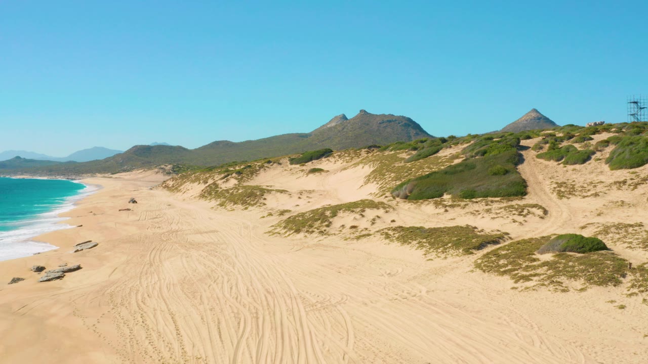 East Cape, Baja California Sur aerial view with the beach stretching diagonally, a small vehicle near the sand tracks, turquoise water at left, dunes at right, and no visible people.