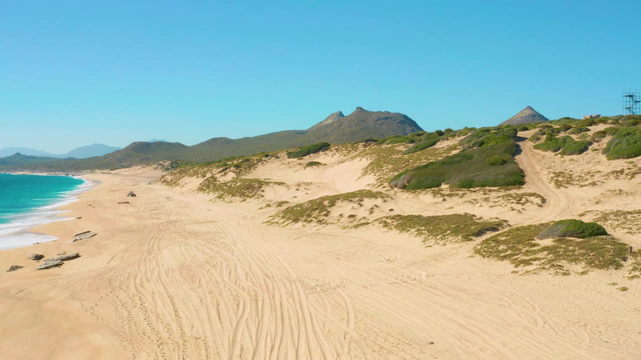 Aerial frame over East Cape, Baja California Sur showing the vehicle farther along the sandy beach, surf curling at left, dune slopes at right, and no visible people.