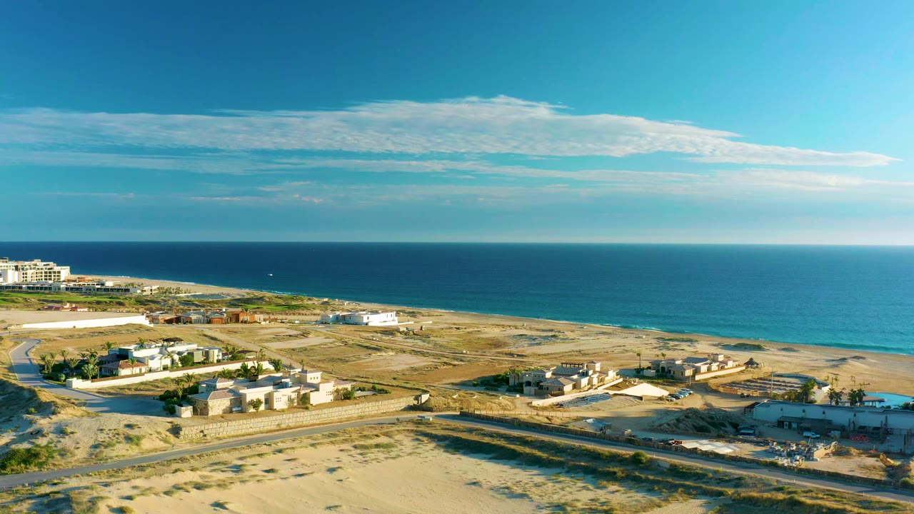 Wide drone view of sandy lots, a coastal road, buildings, and ocean beyond.