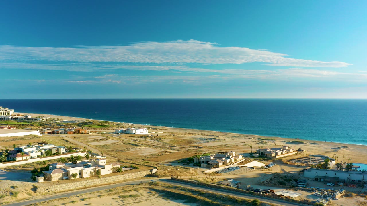 Aerial frame over Diamante Cabo San Lucas showing a road, sandy lots, low buildings, blue ocean, scattered clouds, and no visible people.
