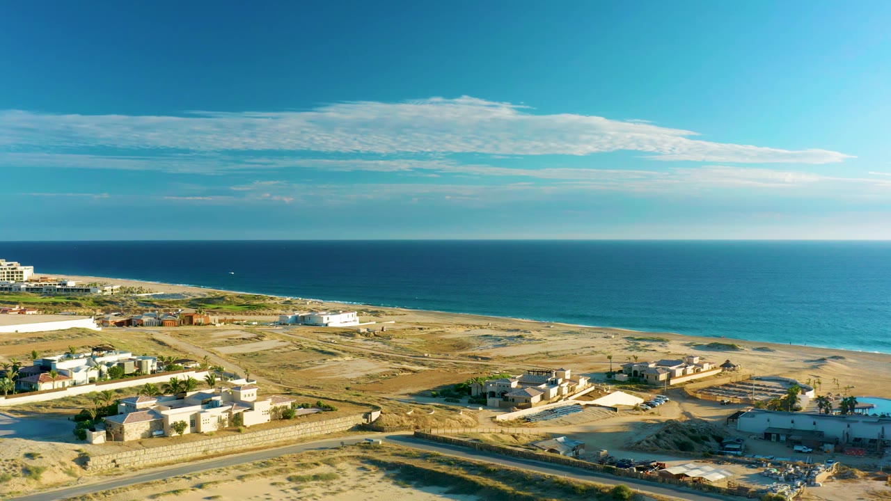 Aerial view in Diamante Cabo San Lucas with white low buildings at left, open sandy development land, calm blue water, a long cloud band, and no visible people.