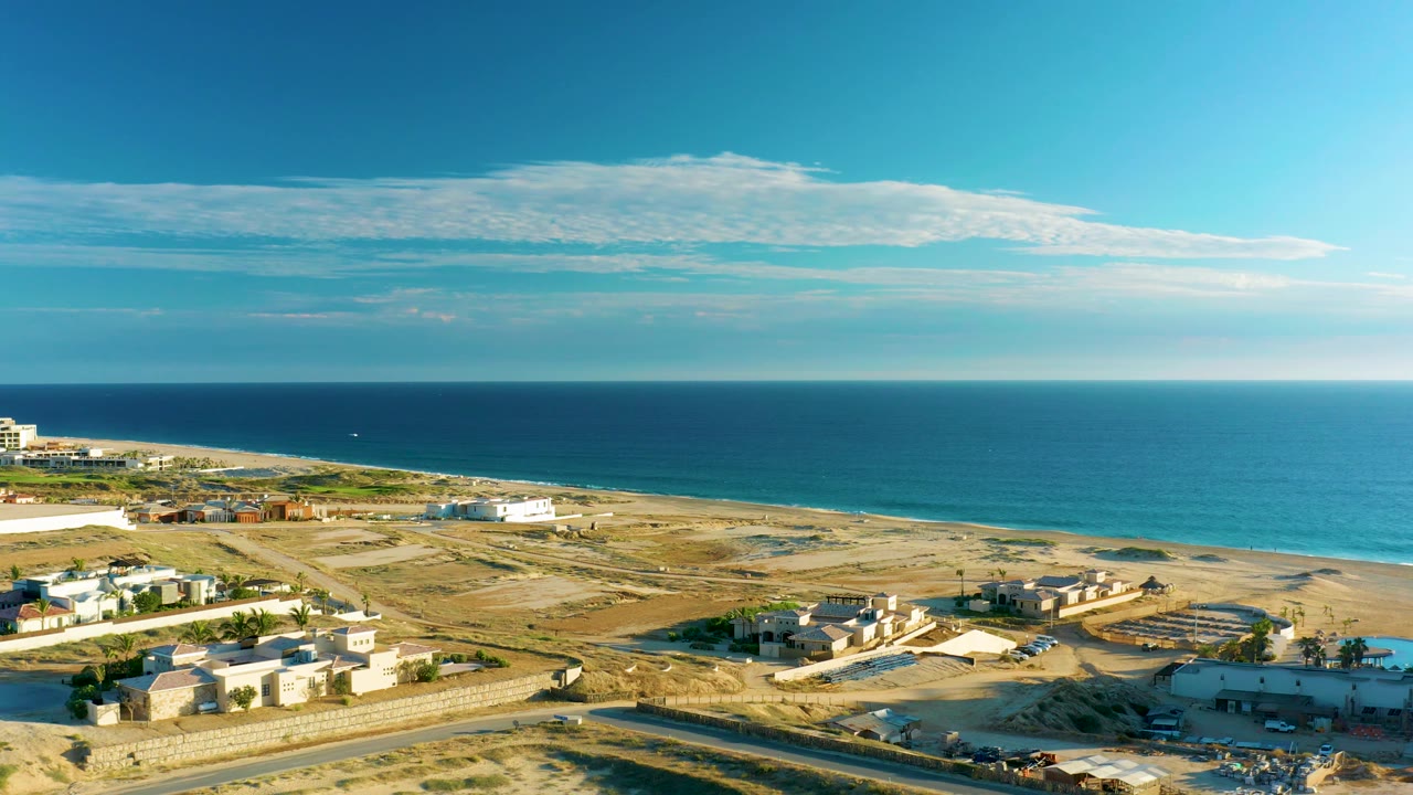 Drone frame over Diamante Cabo San Lucas showing more sandy open ground, scattered coastal buildings, the ocean stretching across the background, and no visible people.