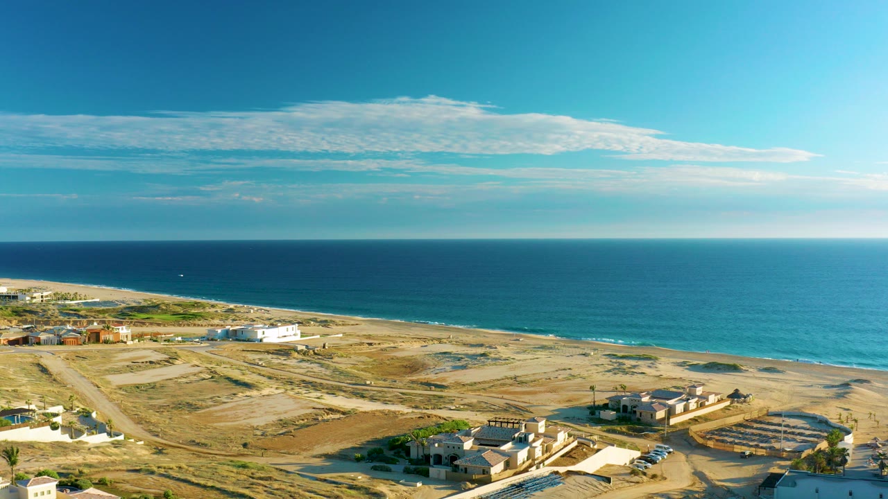 Aerial frame over Diamante Cabo San Lucas with sandy graded lots, scattered coastal buildings, a beach strip, deep blue ocean, long clouds, and no visible people.
