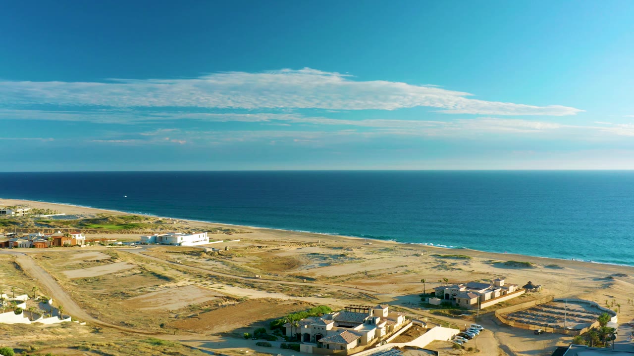 Drone frame over Diamante Cabo San Lucas showing tan construction-like lots, low homes or structures, a narrow beach, blue ocean, bright sky, and no visible people.