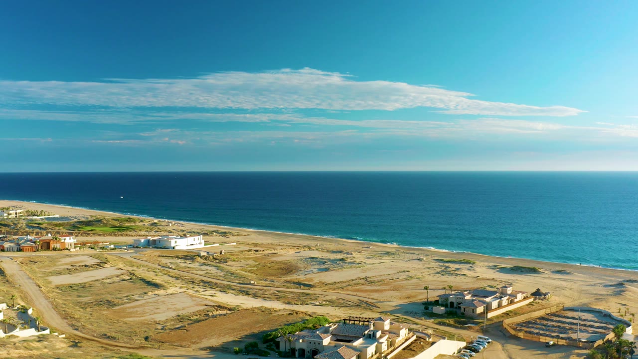 Wide aerial of ocean, beach, road, and sparse coastal buildings under clear sky.