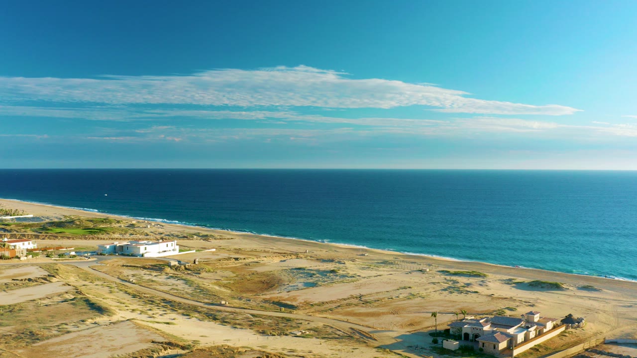 Drone frame over Diamante Cabo San Lucas showing sandy dunes, a low white beachfront structure, turquoise ocean, blue sky, thin clouds, and no visible people.