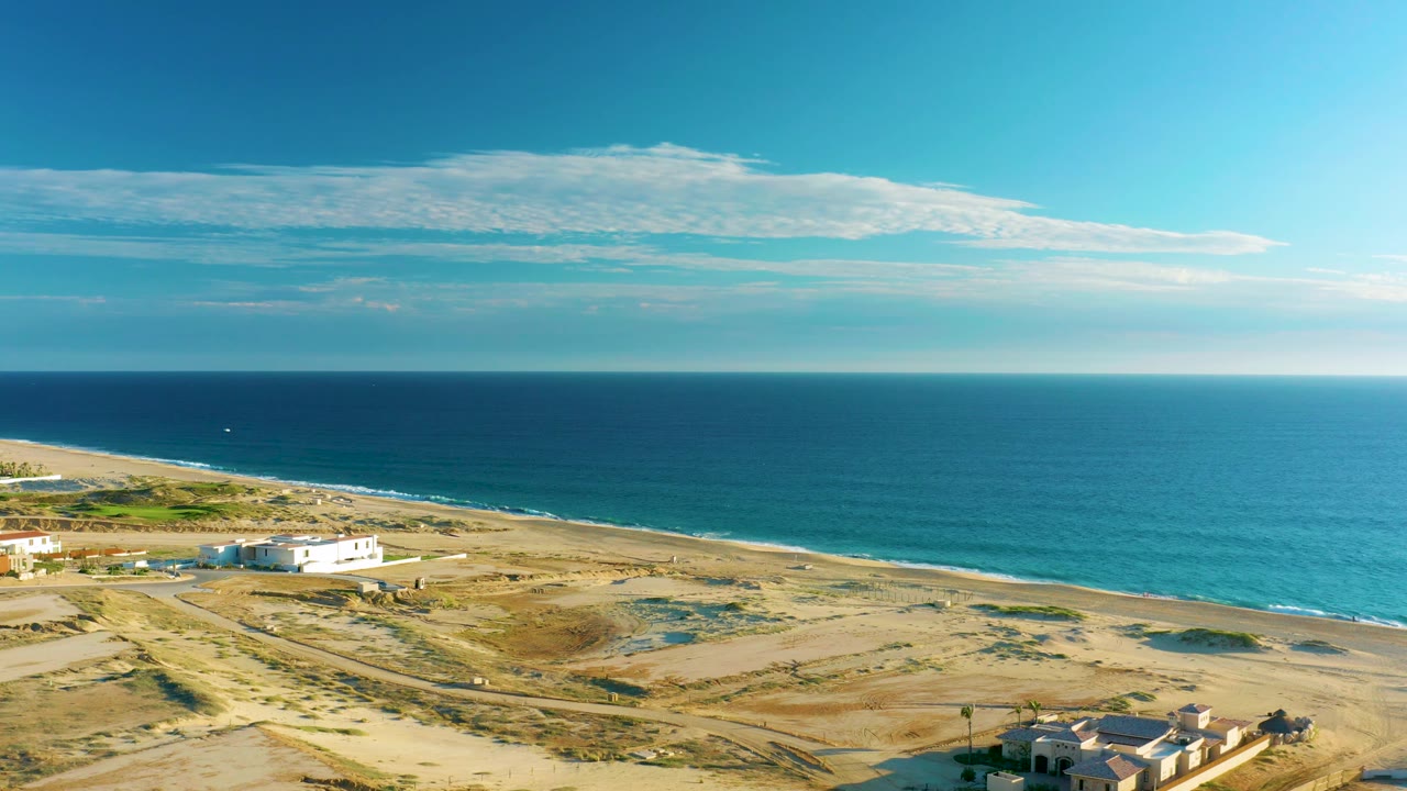 Diamante Cabo San Lucas aerial frame showing beachfront structures near the lower right, open dunes across the foreground, ocean beyond the beach, and no visible people.