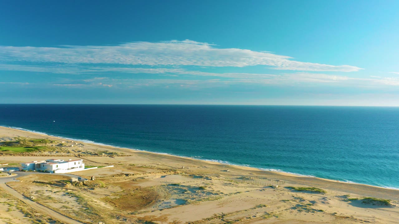 At 0:00, an aerial view shows a white coastal house on sandy terrain beside the beach and blue ocean in Diamante Cabo San Lucas, Mexico, with no visible people.