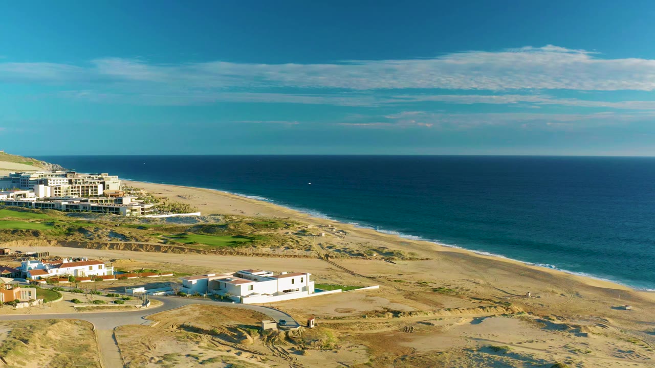 Drone view in Diamante Cabo San Lucas showing a curving road, white buildings, sandy dunes, a broad beach, blue water, and no visible people.