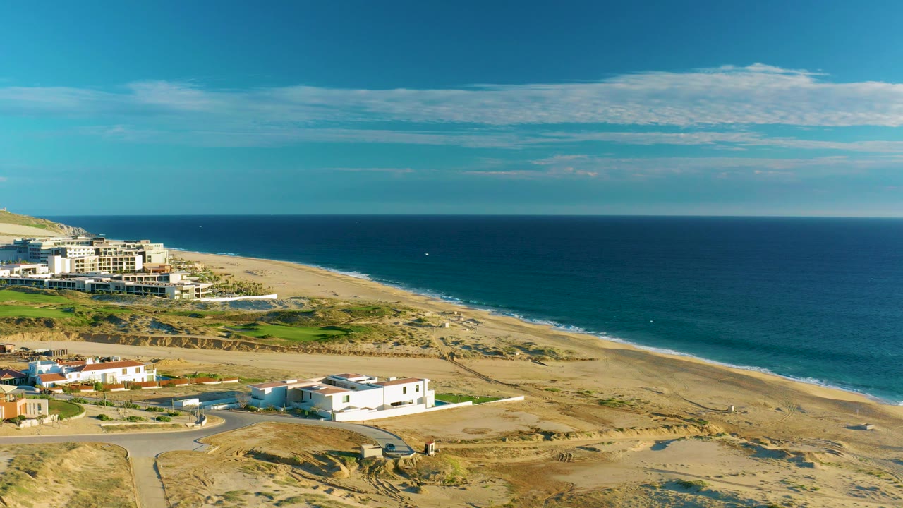 Aerial coastline with road, white buildings, dunes, and deep blue water.