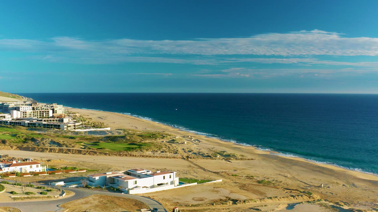 Aerial frame over Diamante Cabo San Lucas showing white coastal buildings in the lower foreground, a long sandy beach, blue ocean, scattered clouds, and no visible people.
