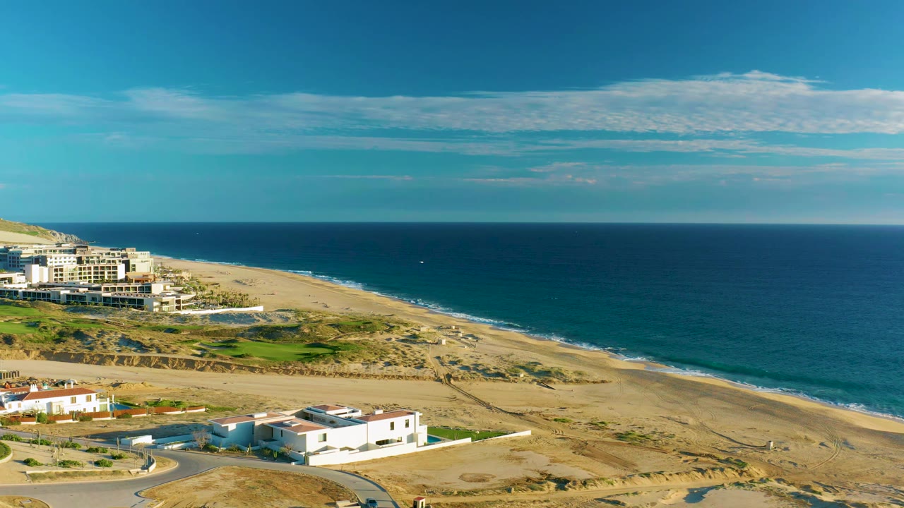Drone view over Diamante Cabo San Lucas with the building cluster shifted farther left, sandy shoreline running diagonally, deep blue water, a small white speck offshore, and no visible people.