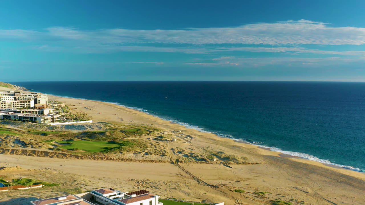 Aerial view at Diamante Cabo San Lucas showing the beach running diagonally beside calm blue ocean, with resort buildings and green managed grass on the left and no visible people.