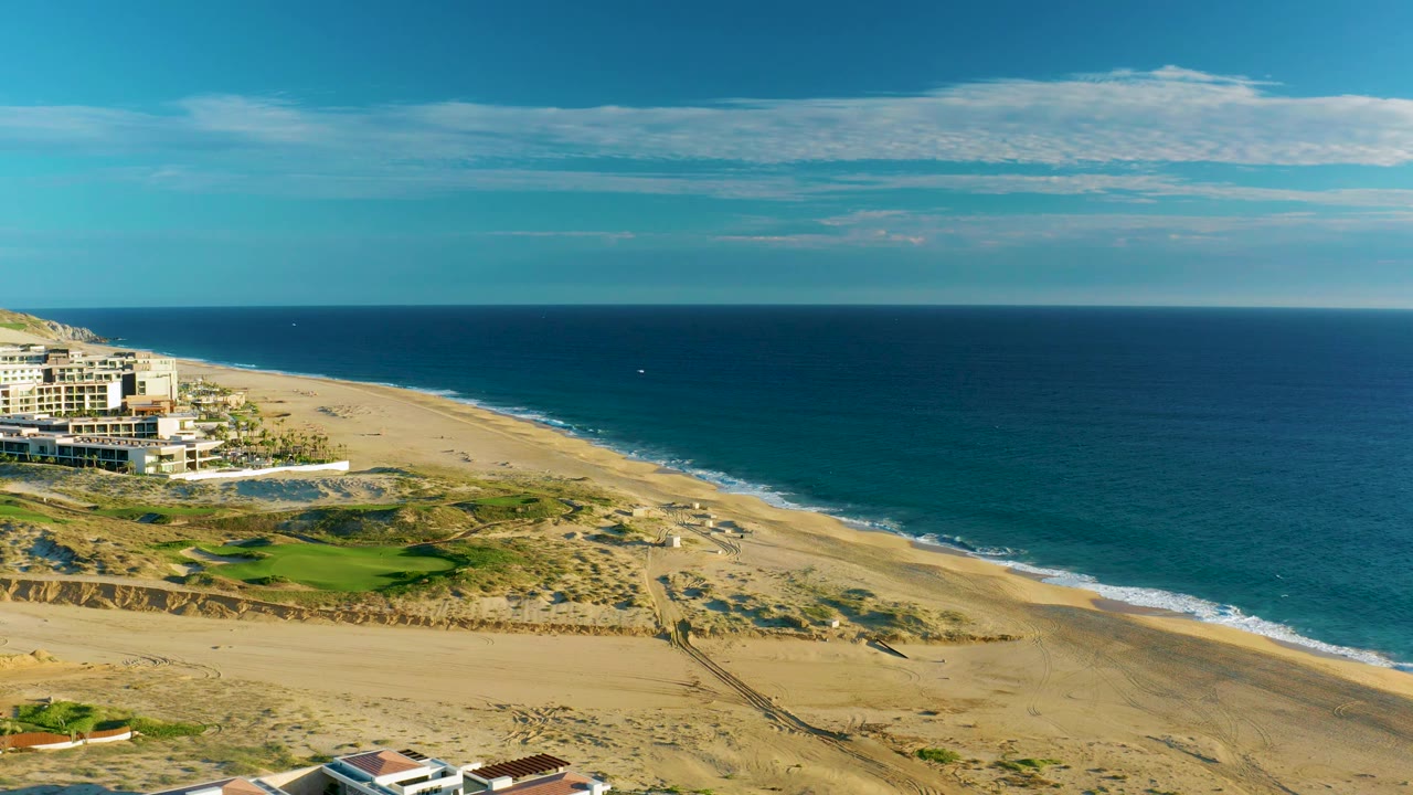 Mid-clip aerial frame over Diamante Cabo San Lucas with a broad tan beach, dark blue ocean, low white waves, green grass near dunes, left-side buildings, and no visible people.