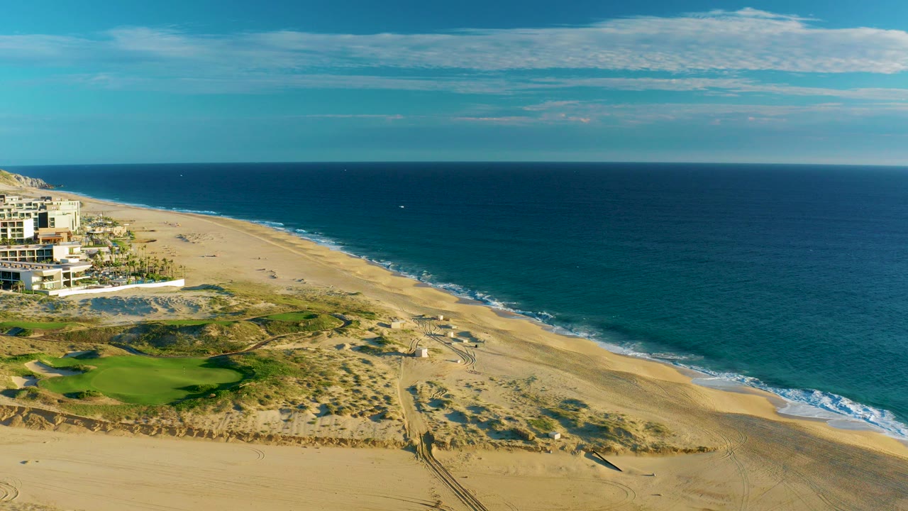Aerial view over Diamante Cabo San Lucas with a sandy beach curving beside blue ocean, green golf course areas at lower left, coastal buildings on the far left, and no visible people.