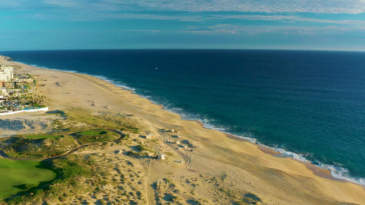 High aerial view over Diamante Cabo San Lucas showing tan beach, blue ocean, dune tracks, green landscaped ground, coastal buildings at left, and tiny distant beachgoers.