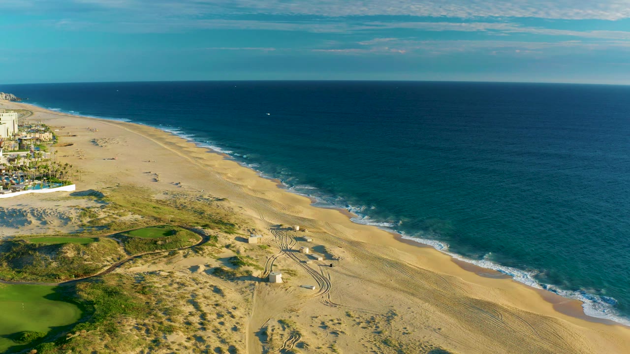 Drone-style aerial over Diamante Cabo San Lucas with turquoise nearshore water, pale sand, dune vegetation, curved green landscaping, coastal structures at left, and small distant beach figures.