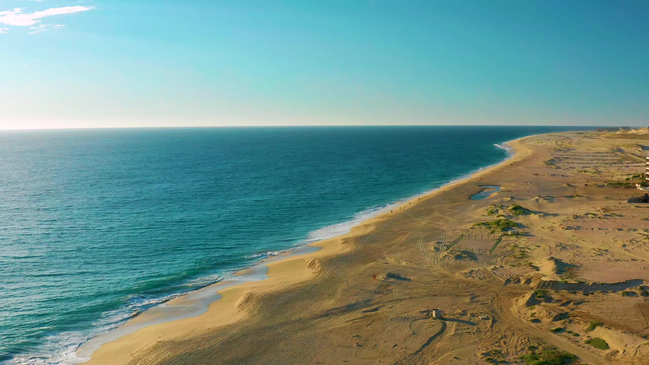 Aerial frame over Diamante Cabo San Lucas showing blue ocean on the left, a long sandy beach curving into dry dunes on the right, clear sky above, and no visible people.