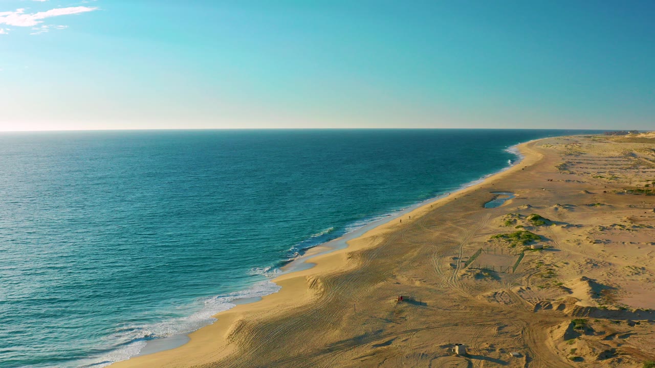 Aerial view over Diamante Cabo San Lucas with turquoise ocean at left, a sandy beach curving into the distance, dune tracks inland, and tiny people and a vehicle visible on the sand.