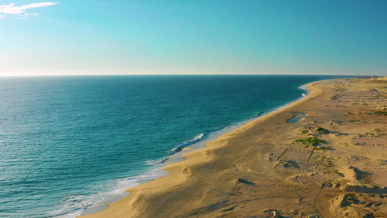 Aerial coastline view in Diamante Cabo San Lucas with the ocean filling the left side, a narrow surf band, sandy dunes to the right, a small vehicle on the beach, and tiny people visible as dots.