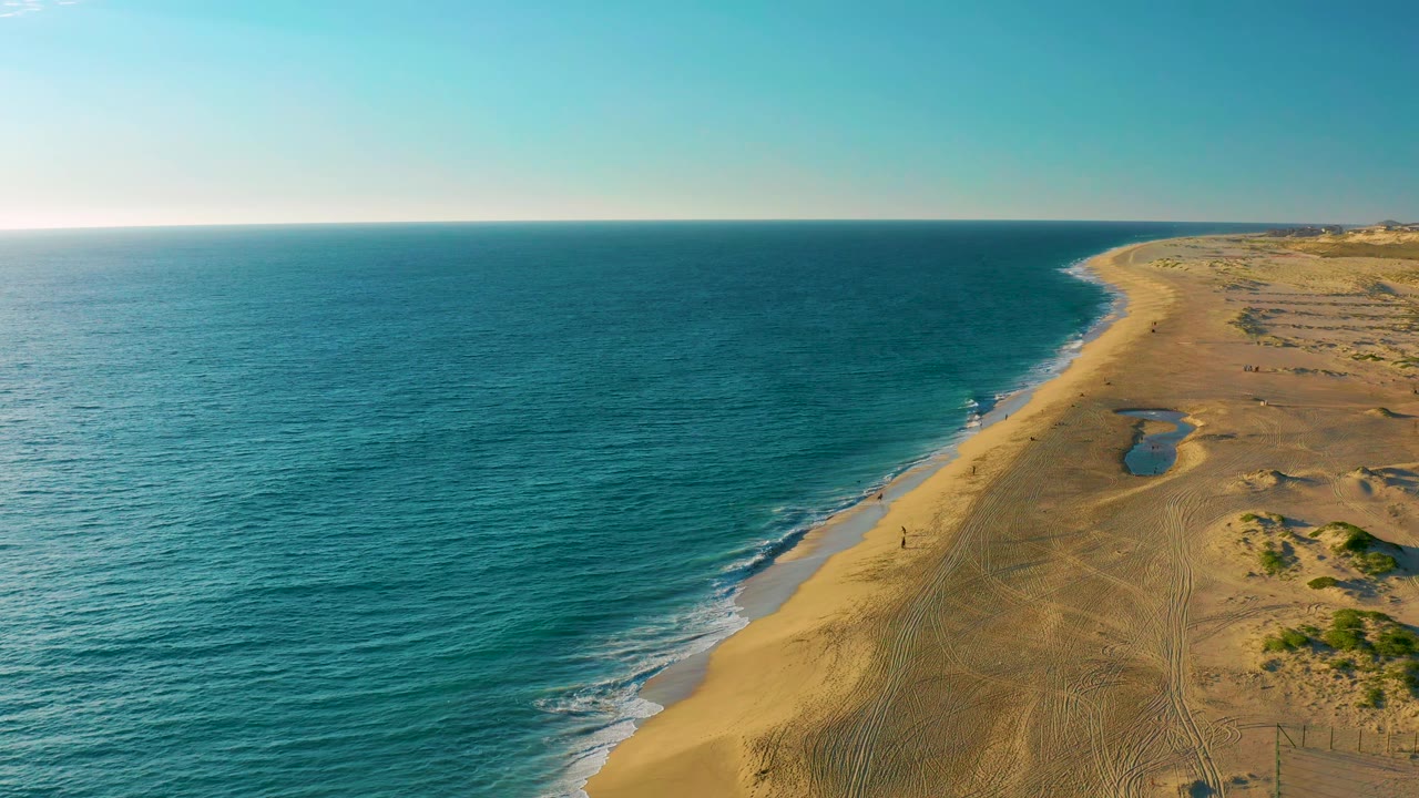 Aerial view in Diamante Cabo San Lucas with gentle white surf tracing the beach, open ocean across the left half, sandy dunes to the right, and no visible people.