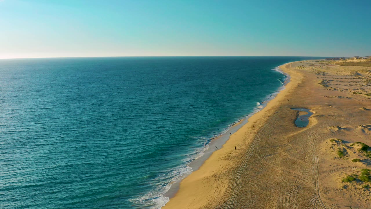 Drone-height view of the Diamante Cabo San Lucas beach with rippled blue-green water, a narrow line of foam along the sand, dune tracks on the right, and no visible people.